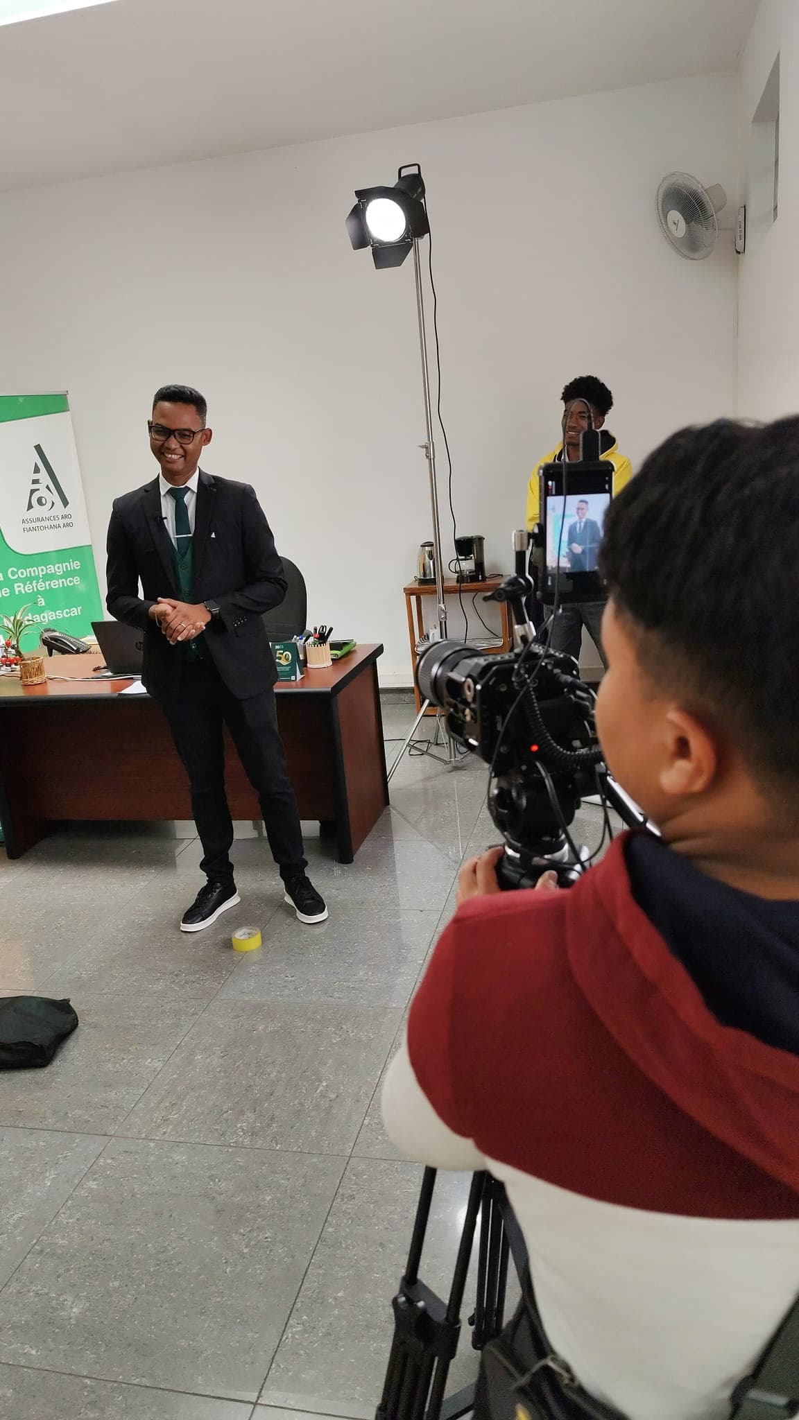 Tournage d’un témoignage dans un bureau d’agence ARO : l’intervenant devant un kakémono de la compagnie, un projecteur sur pied, un opérateur caméra au premier plan et un second technicien en arrière-plan